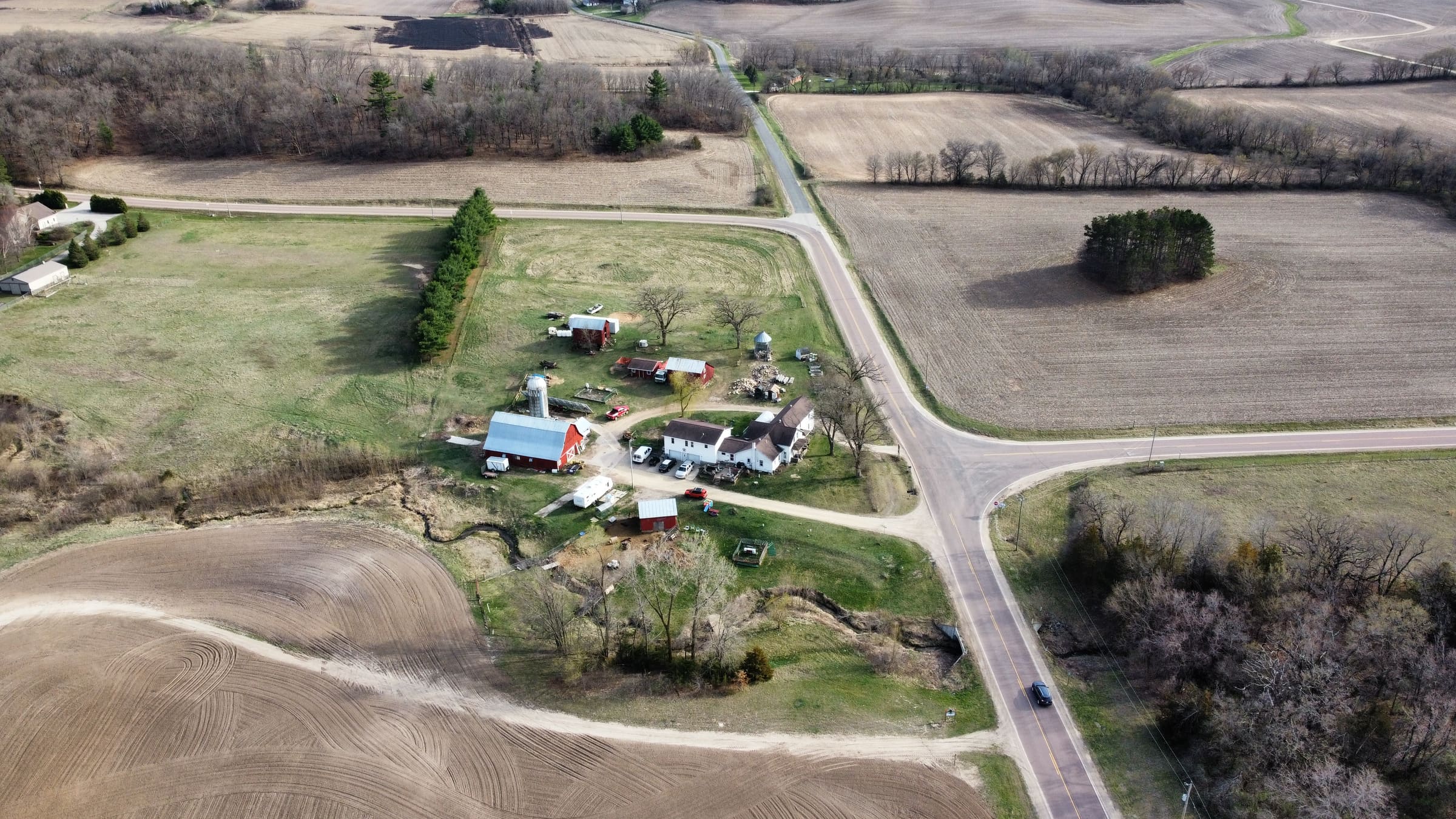Aerial view of a farmstead with a red barn and white farmhouse at a rural crossroads, surrounded by freshly tilled fields.